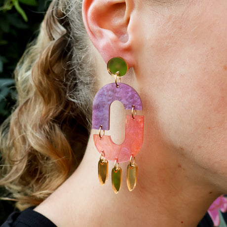 Close-up of a person wearing statement oval shaped purple pink and gold dangle earrings