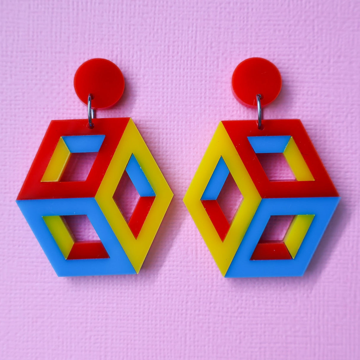 Colourful red, blue and yellow statement geometric cube earrings on a pink background