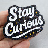 black Glittery 'Stay Curious' brooch with white text and yellow stars held in a hand against a neutral background