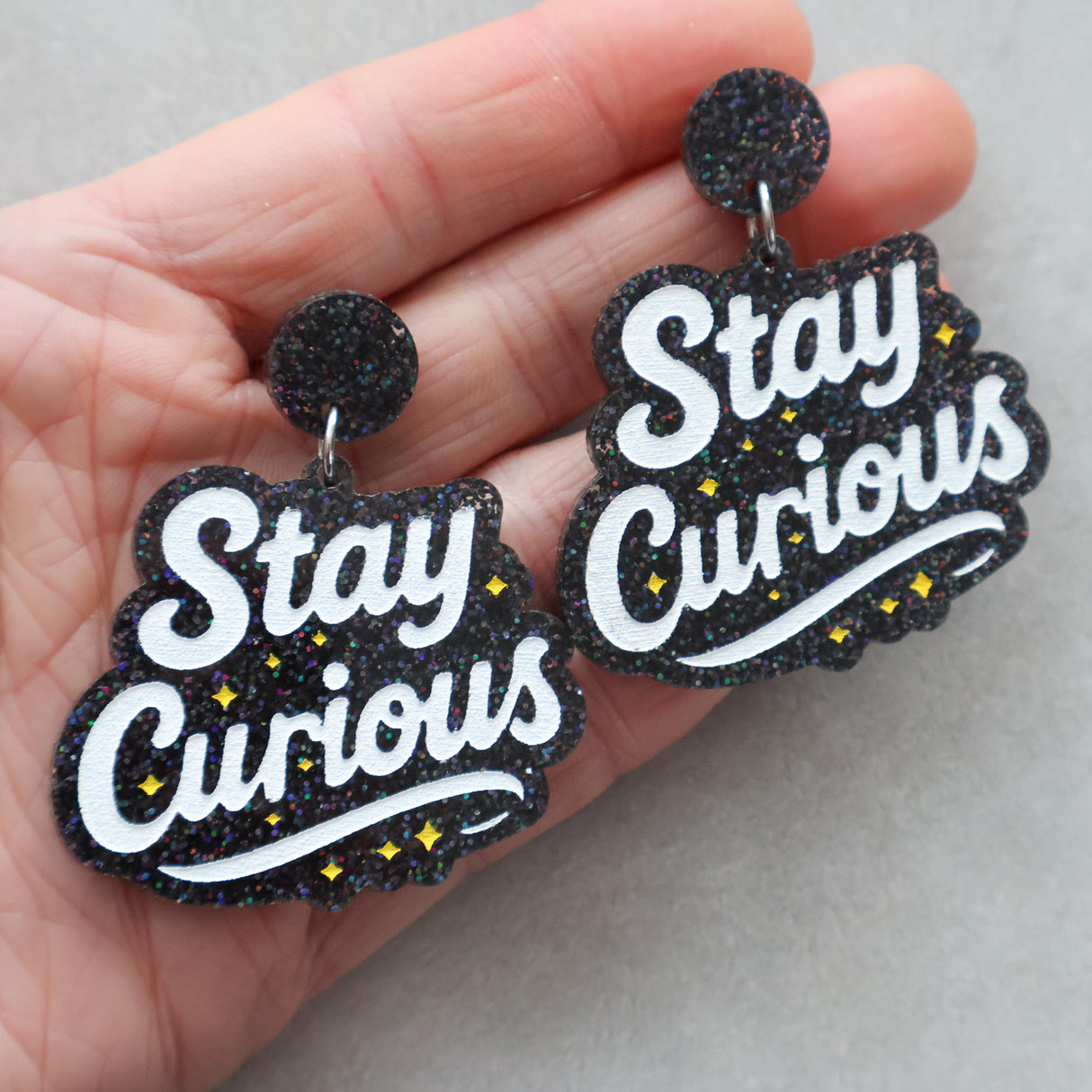 Black glitter earrings with 'Stay Curious' text held in a hand