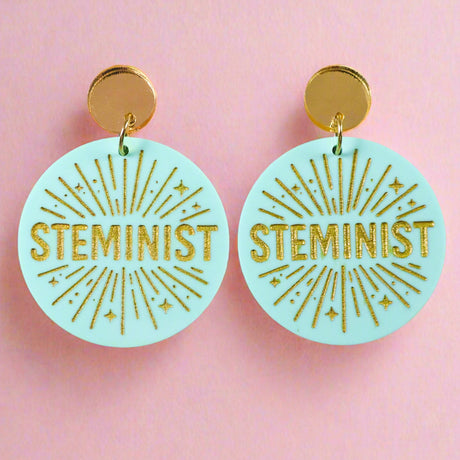 Teal and gold toned acrylic earrings with 'STEMINIST' text on a pink background