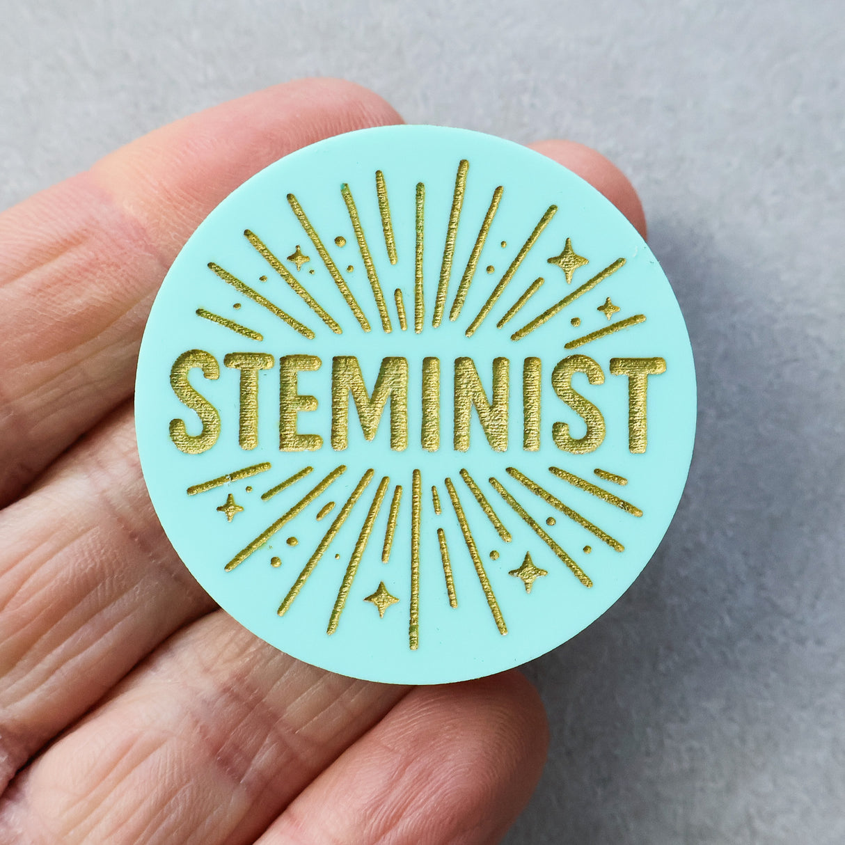 Round teal acrylic brooch with 'STEMINIST' in gold text held in a hand