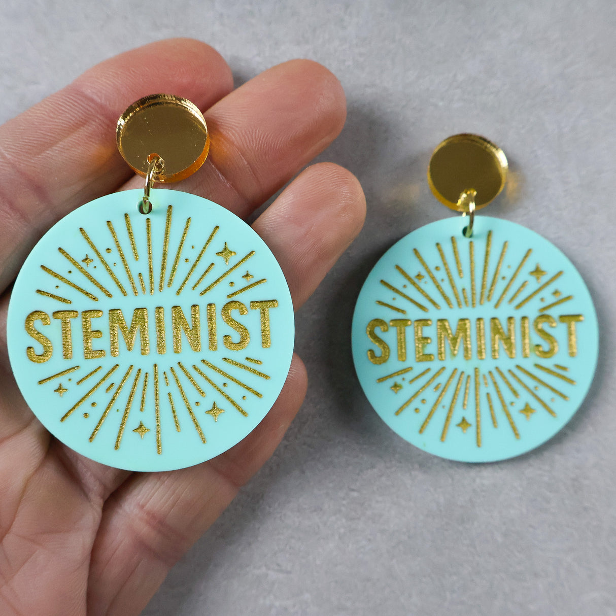 Turquoise or teal dangle earrings with 'STEMINIST' gold text and gold mirror acrylic toppers held by a hand on a gray background