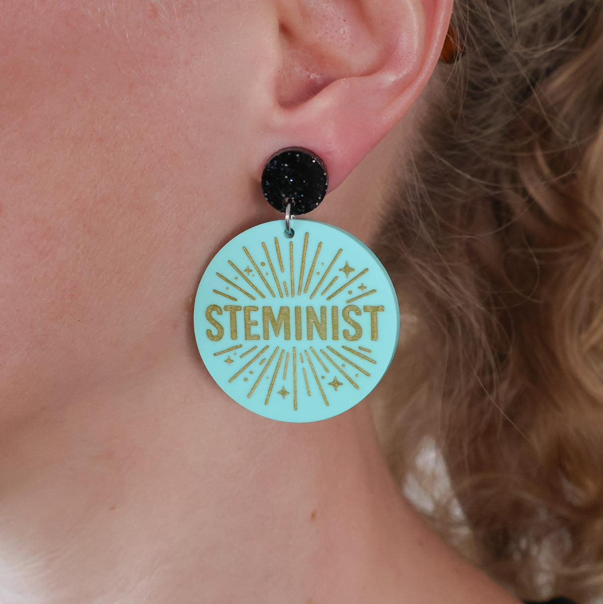 Earring with 'STEMINIST' text on a light teal acrylic being modelled on an ear