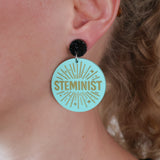 Earring with 'STEMINIST' text on a light teal acrylic being modelled on an ear