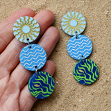 Colourful acrylic disc dangle earrings held in a hand with a sun, waves and seaweed/fish graphic. 
