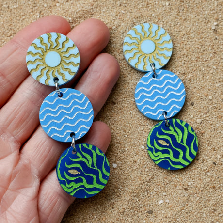 Colourful acrylic disc dangle earrings held in a hand with a sun, waves and seaweed/fish graphic. 