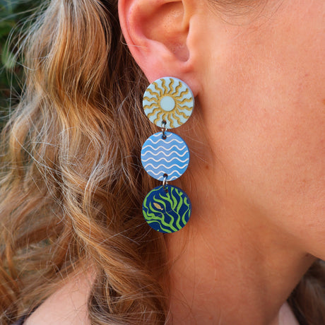 Close-up of a person wearing colorful earrings with sun waves and seaweed patterns.