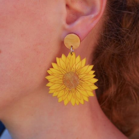 A close-up of a person's ear wearing a yellow daisy-shaped drop earring with a round topper.