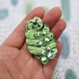Green acrylic laser cut tardigrade brooch in hand for scale. Brooch has black and white handpainted details.