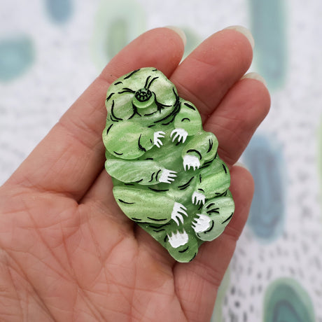 Green acrylic laser cut tardigrade brooch in hand for scale. Brooch has black and white handpainted details.