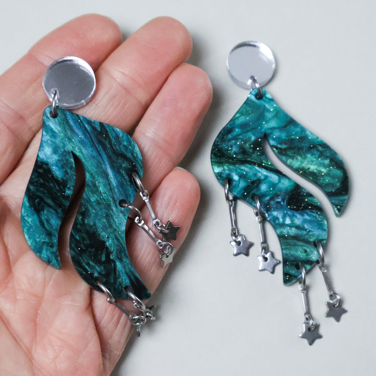Teal wave shaped marbled acrylic earrings with star charms held by a hand, with silver mirror acrylic toppers. 