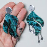 Teal wave shaped marbled acrylic earrings with star charms held by a hand, with silver mirror acrylic toppers. 