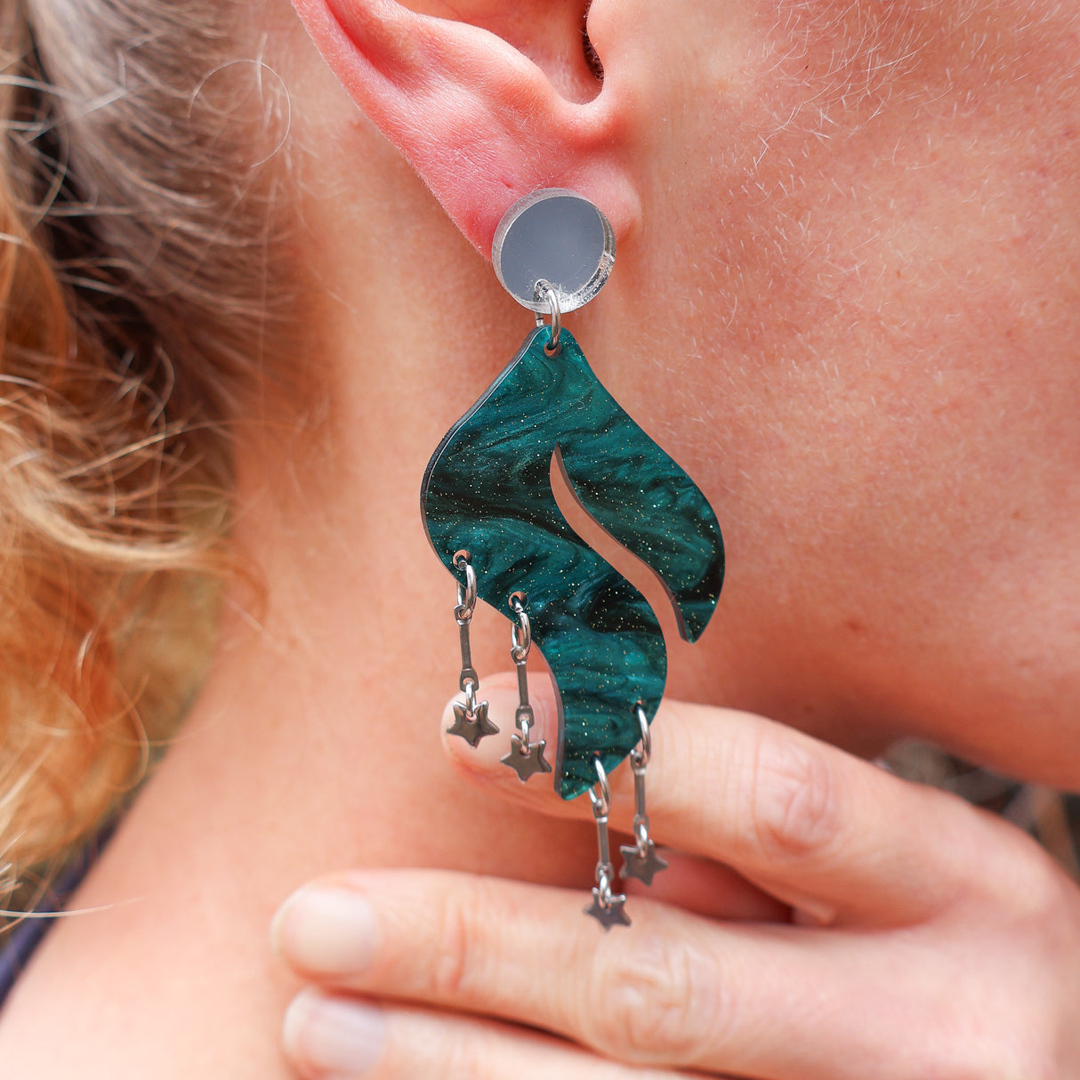 Close-up of a person wearing a green feather or wave shaped earring with star charms.
