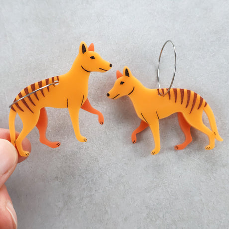 Two orange thylacine or tasmanian tiger shaped earrings with round stainless steel hoops. One earring is being held between fingers. 
