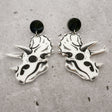 A pair of triceratops skull acrylic earrings in black and white with round black acrylic toppers against a grey background. 