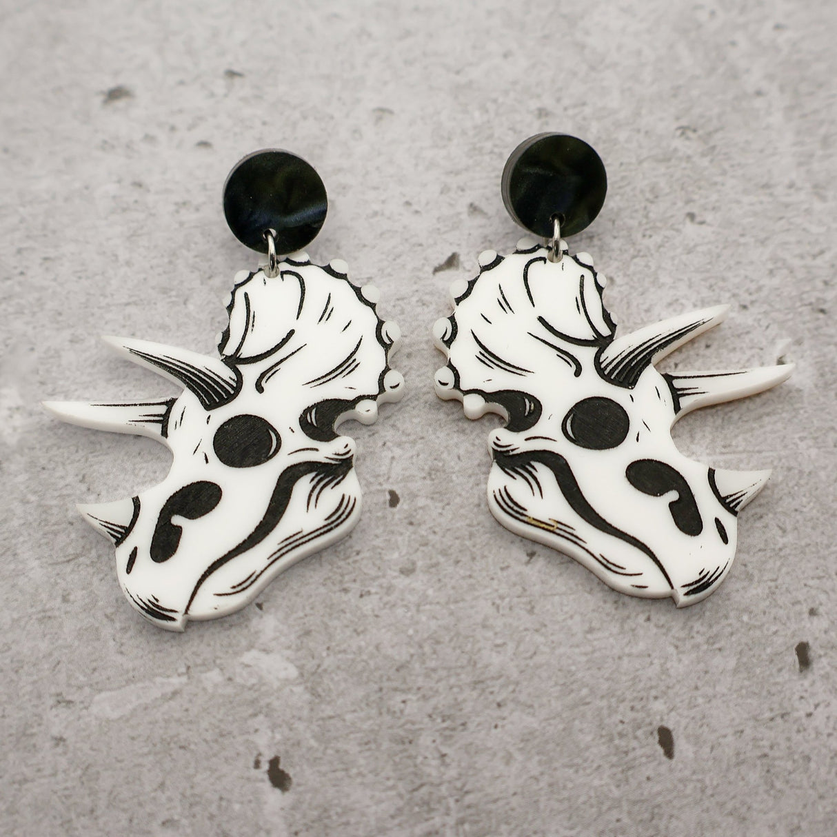 A pair of triceratops skull acrylic earrings in black and white with round black acrylic toppers against a grey background. 
