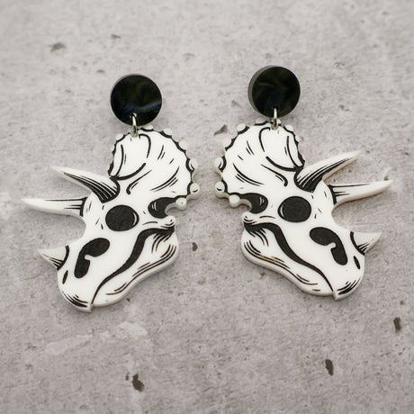 A pair of triceratops skull acrylic earrings in black and white with round black acrylic toppers against a grey background. 