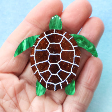 Hand holding a small turtleacrylic brooch with a brown shell and green limbs against a light blue background.