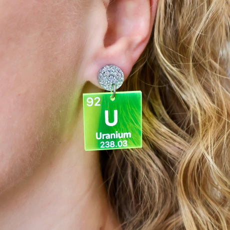 Uranium periodic table earrings, with white text on bright yellow transparent acrylic squares, hanging from silver glitter toppers. Earrings being modelled.