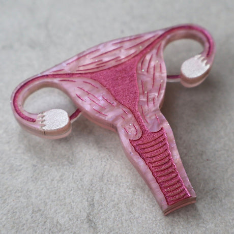 A closeup view of a laser cut acrylic uterus brooch, in pink acrylic with pink painted details. 