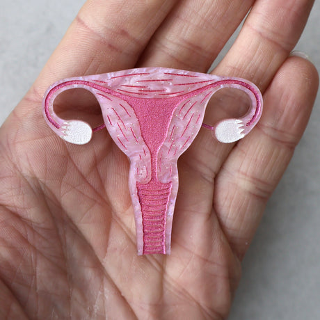 A closeup view of a laser cut acrylic uterus brooch, in pink acrylic with pink painted details being held in a pam.