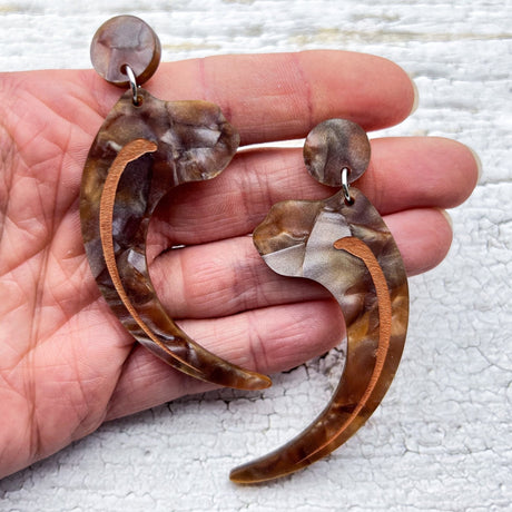 Velociraptor claw earrings, laser cut from textured brown acrylic, and hanging from brown acrylic earring toppers. Being held in hand to show scale.
