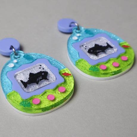 A pair of colourful earrings shaped like a virtual pet gaame, displaying sharks. 