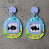 closeup view of a pair of colourful earrings shaped like a virtual pet gaame, displaying sharks. 