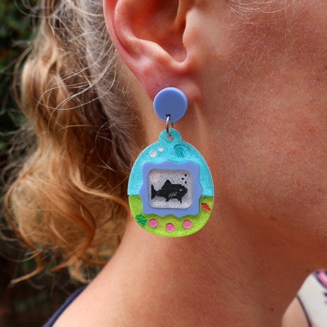 Close-up of a person wearing a colorful earring shaped like a virtual pet game with a shark design.