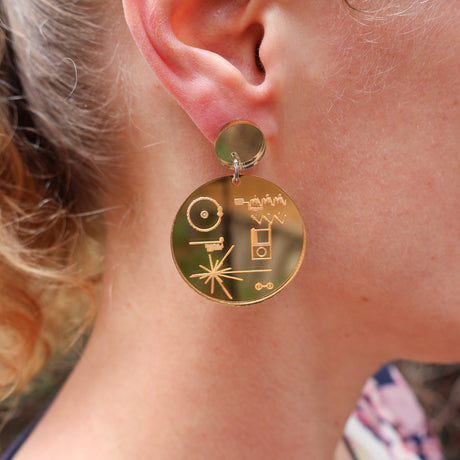 Close-up of a person wearing gold mirror acrylic earrings dangle with voyager golden record designs.