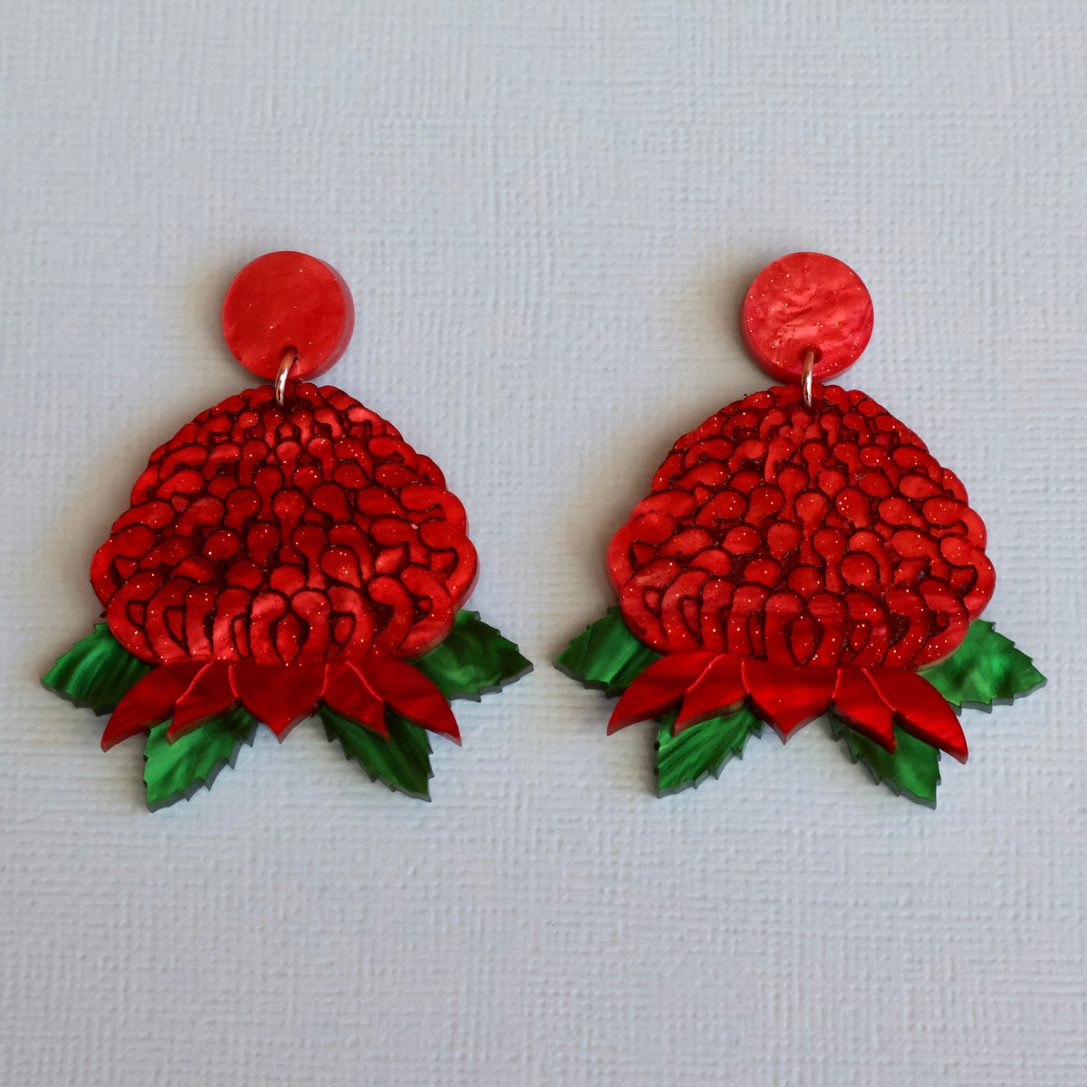 A pair of handmade red and green floral drop waratah earrings on a textured light blue background.