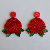 A pair of handmade red and green floral drop waratah earrings on a textured light blue background.