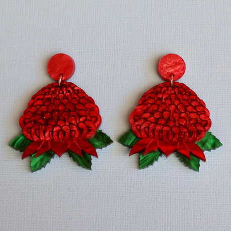 A pair of handmade red and green floral drop waratah earrings on a textured light blue background.