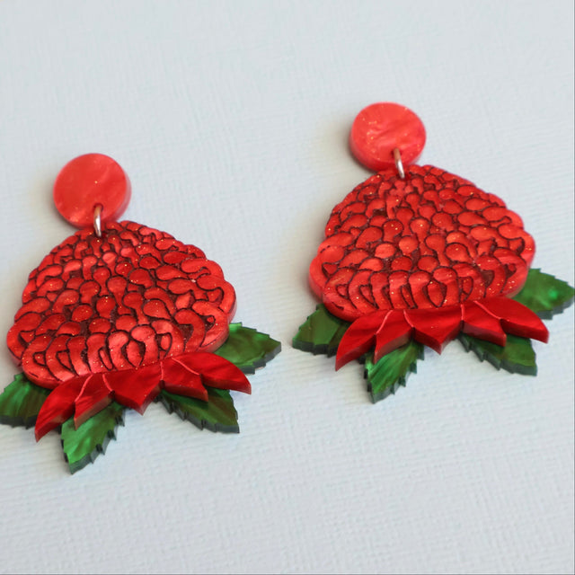 A pair of handmade red waratah-shaped earrings with green leaf accents, displayed on a light textured surface.