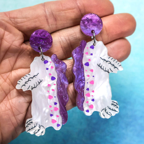Closeup of laser cut acrylic white and purple nudibranch earrings with purple toppers being held in hand.
