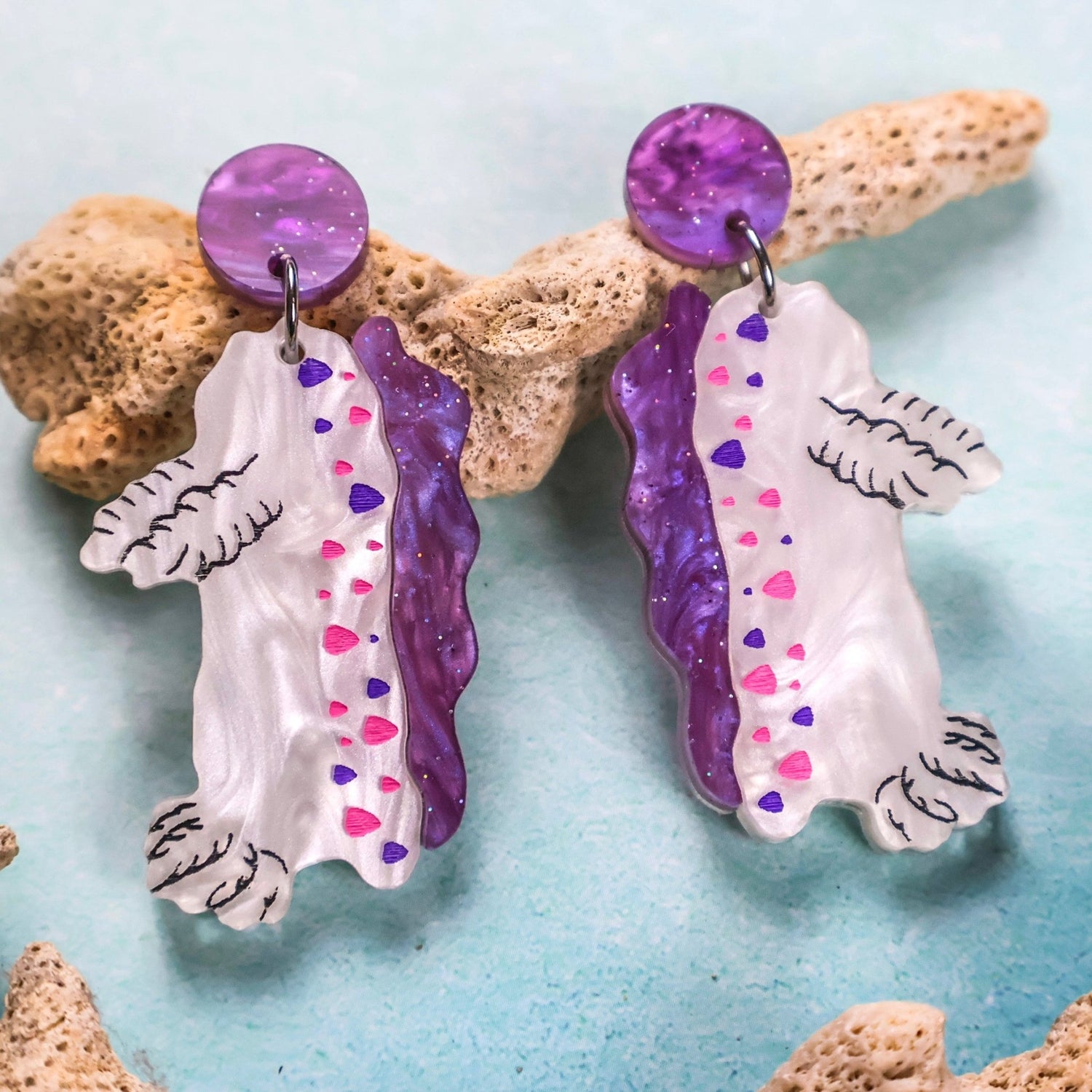 Pair of acrylic nudibranch or sea slug earrings with purple and white design on a light blue background with coral. Marine Biology Science Earrings