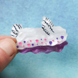 Small white pink and purple nudibranch brooch being held in a hand against a blue backdrop