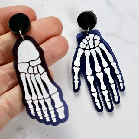 Skeletal hand and foot earrings on a white background with hand holding foot earring