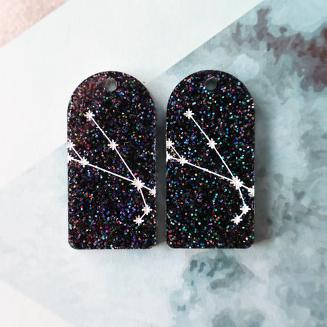 Pair of arch acrylic earrings with aries star constellation design in white again black glitter acrylic. 