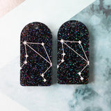 Pair of arch acrylic earrings with libra star constellation design in white again black glitter acrylic.
