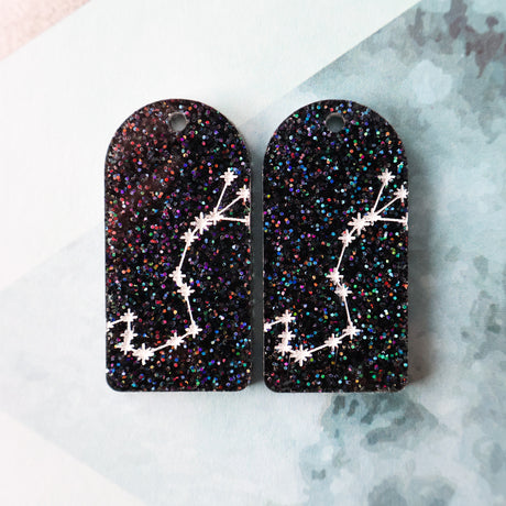 Pair of arch acrylic earrings with scorpio star constellation design in white again black glitter acrylic.