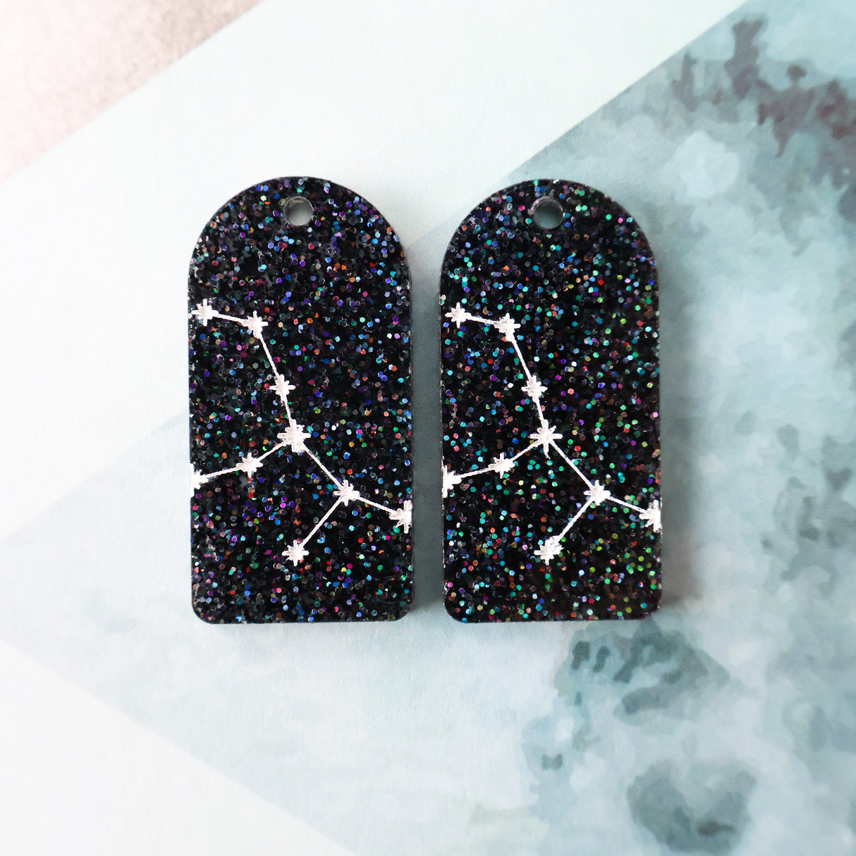 Pair of arch acrylic earrings with virgo star constellation design in white again black glitter acrylic.