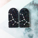 Pair of arch acrylic earrings with virgo star constellation design in white again black glitter acrylic.