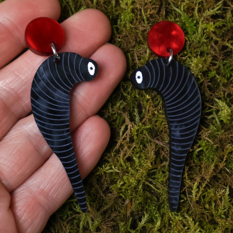 Black leech shaped earrings with red circular toppers held between fingers on a mossy background