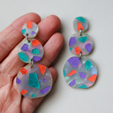 Terrazzo dangle acrylic earrings in orange, purple and teal tones. Dangles are 3 components and statements sized. One side is being held up in hand