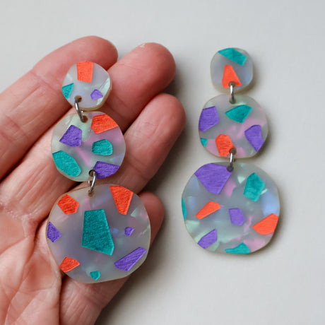 Terrazzo dangle acrylic earrings in orange, purple and teal tones. Dangles are 3 components and statements sized. One side is being held up in hand