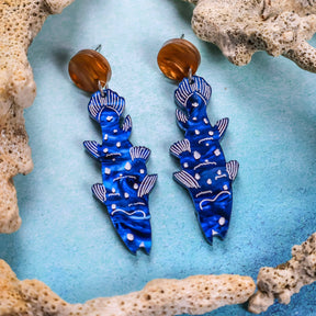A pair of blue acrylic earrings designed to resemble coelacanths, with round brown toppers, against a light blue background