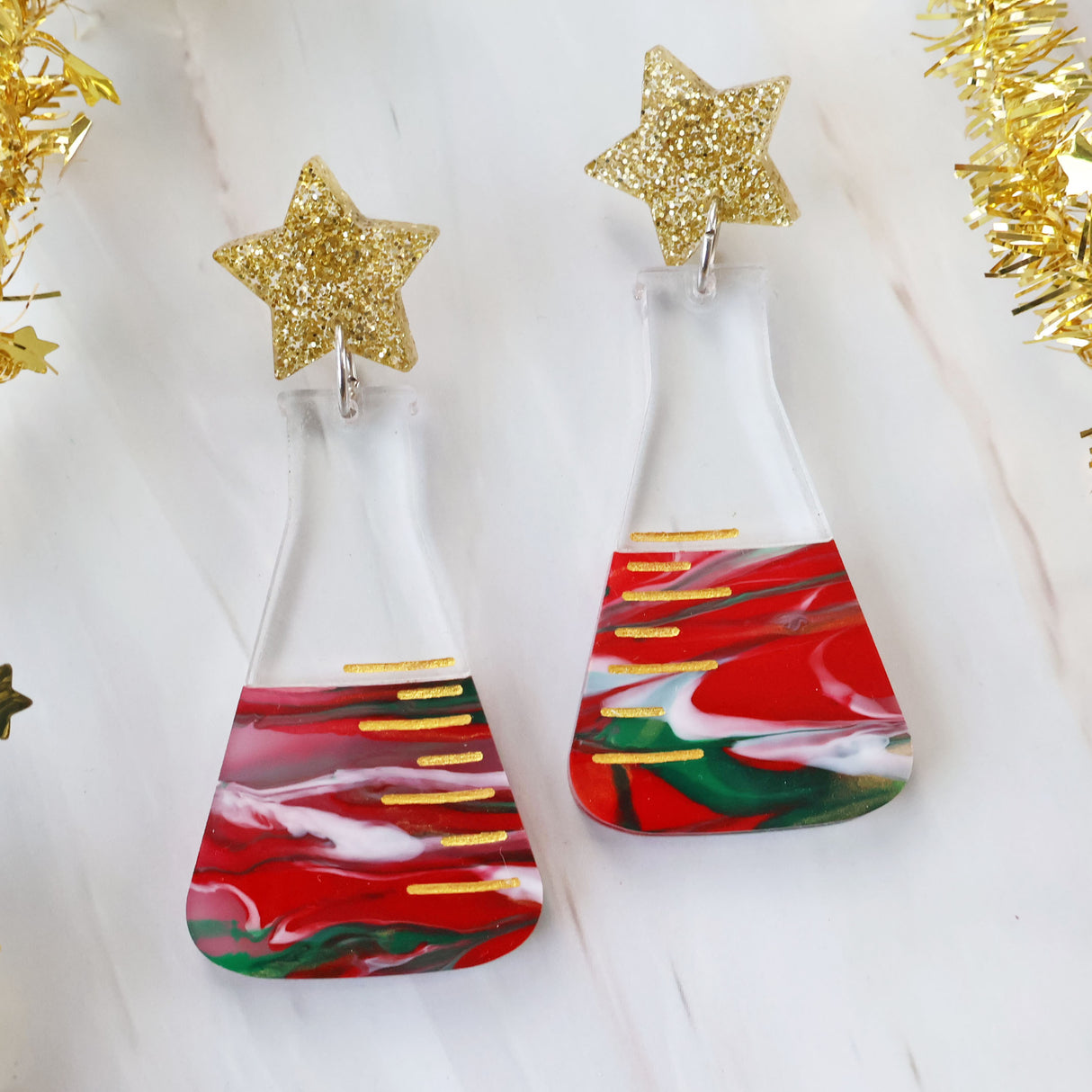 A pair of dangle earrings featuring a conical flask design with Christmas-themed decorations, including red and green swirls and gold accents, topped with glittery star-shaped acrylics.