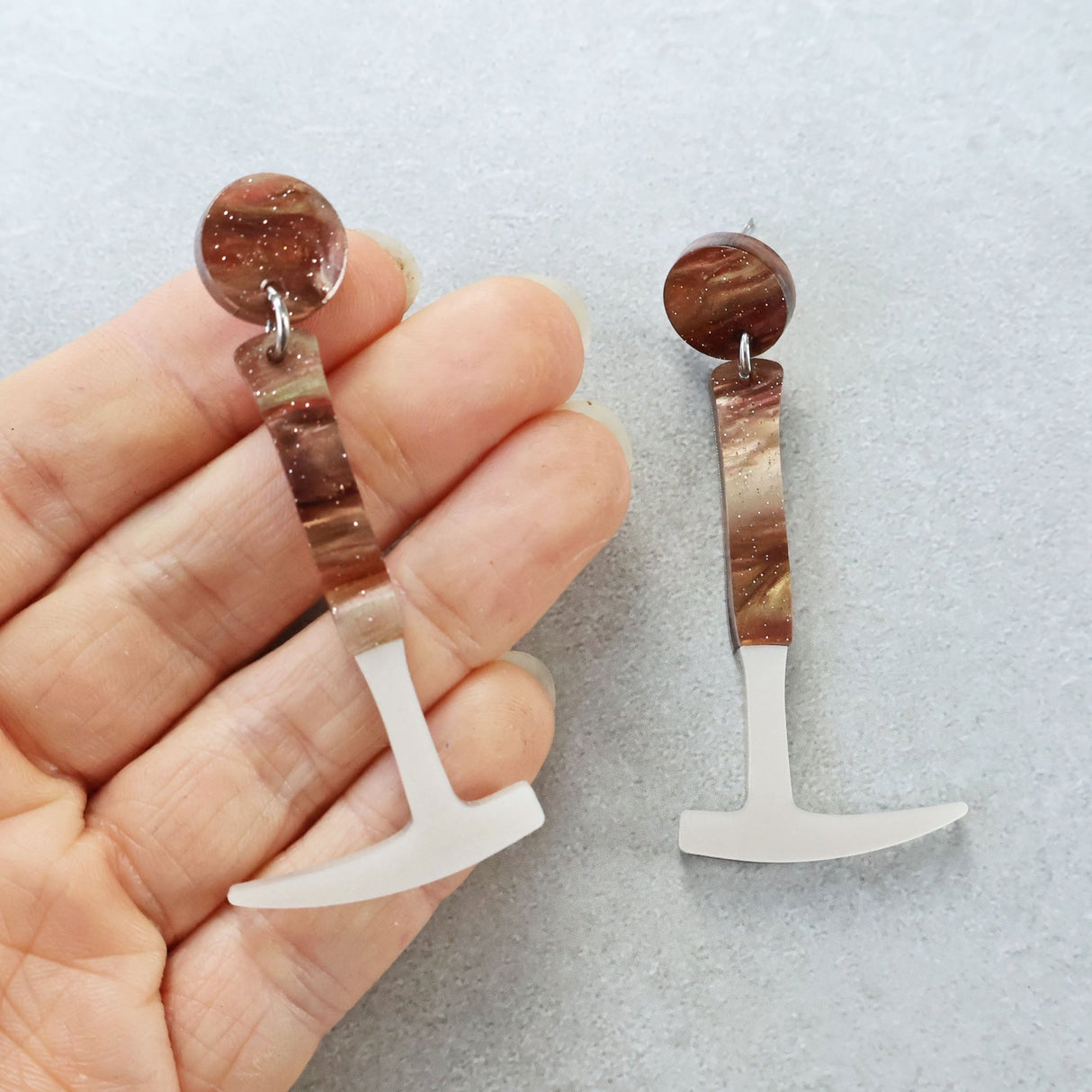 Laser cut acrylic fossil hammer shaped earrings being held up against a grey background. The dangly hammer handle and earring topper has brown swirls and glitter. 